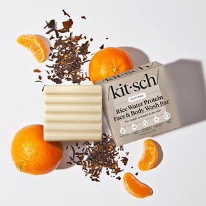 KITSCH Rice Water Protein Restoring Face & Body Wash Bar creative product flat lay — cream ribbed bar and /kit·sch/ packaging surrounded by fresh oranges and loose leaf botanicals — For rough, irritated, & dry skin — Hydrolyzed Rice Protein & Niacinamide Infused