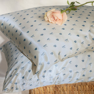 KITSCH Satin Pillowcase in Pearlized Cream & Coastal Blue Print — lifestyle flat lay on wicker surface with pink rose — coastal blue seashells & bows print close-up — Reduces friction against hair & skin, helps reduce breakouts — For hair & skin-loving comfort